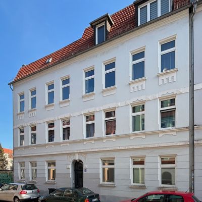 anierter Altbau mit weißer Fassade und rotem Dach in einer ruhigen Stadtstraße mit geparkten Autos.