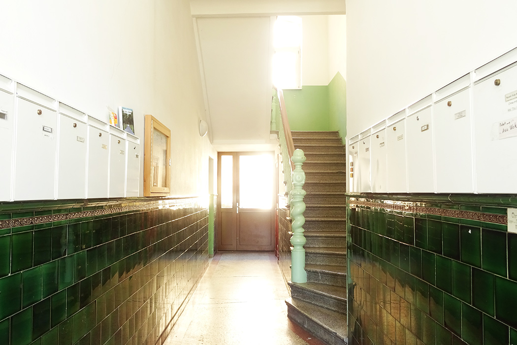 Historischer Hausflur mit grünen Fliesen, weißen Briefkästen und einer klassischen Treppe in einem Altbau mit viel Tageslicht.