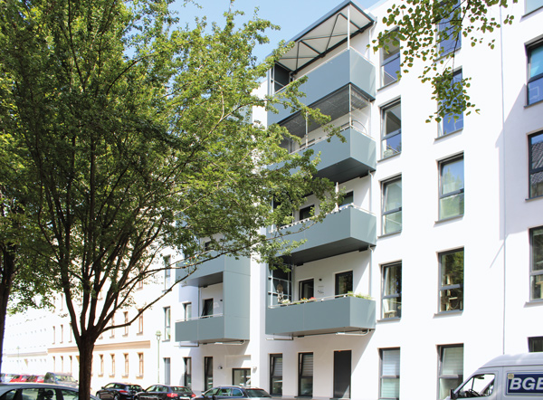 Modernisiertes Wohngebäude mit Balkonen und grünen Bäumen in einer urbanen Umgebung, helle Fassade und gepflegte Architektur.