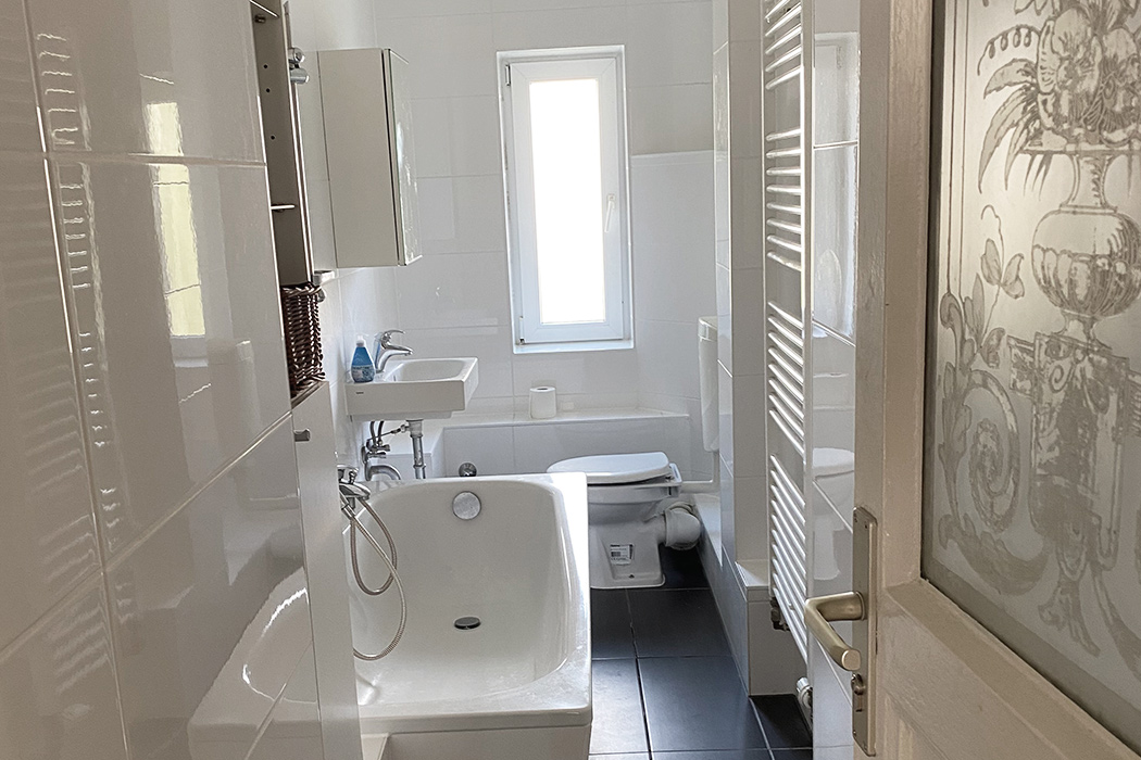 Modernes, helles Badezimmer mit Badewanne, Waschbecken, WC und Fenster, gefliest in Weiß und Schwarz.