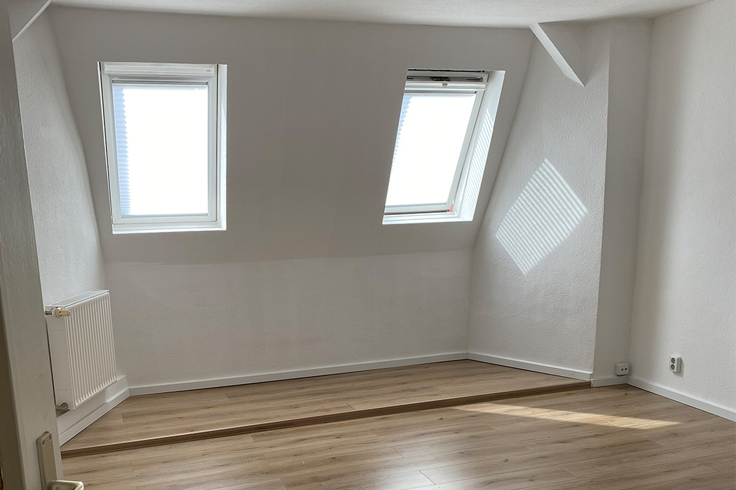 Helles Dachgeschosszimmer mit zwei Fenstern, Laminatboden und weiß gestrichenen Wänden.