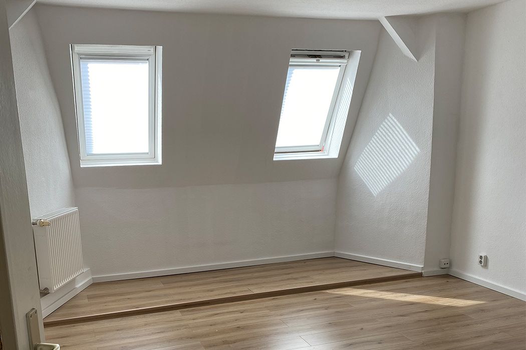 Helles Dachgeschosszimmer mit zwei Fenstern, Laminatboden und weiß gestrichenen Wänden.