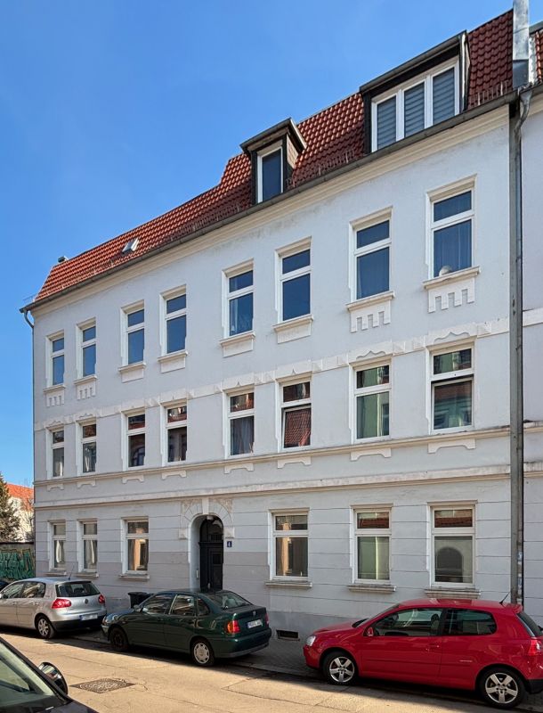 anierter Altbau mit weißer Fassade und rotem Dach in einer ruhigen Stadtstraße mit geparkten Autos.