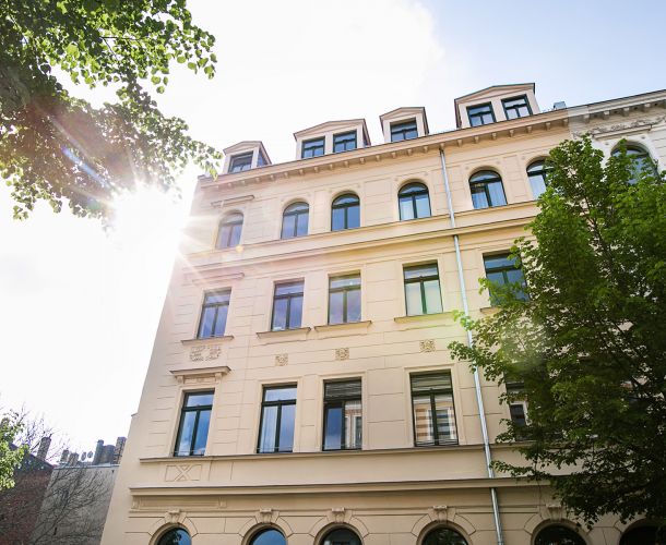 Saniertes Altbau-Gebäude in der Sonne mit grünen Bäumen im Vordergrund.