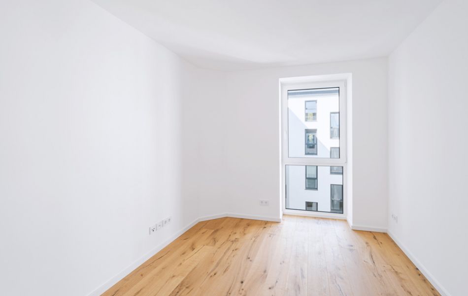 Helles, modernes Zimmer mit Parkettboden, weiß gestrichenen Wänden und bodentiefem Fenster mit Blick auf ein Wohngebäude.