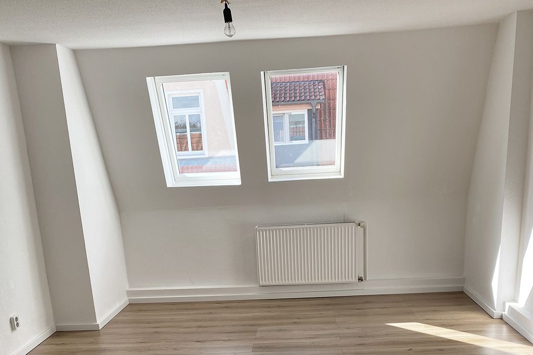 Heller Dachgeschossraum mit zwei schrägen Fenstern, Laminatboden und weiß gestrichenen Wänden, ideal als Wohn- oder Arbeitszimmer.