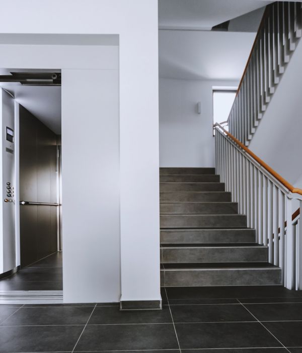 Modernes Treppenhaus mit Aufzug, dunklen Fliesen und hellem Handlauf, barrierefreier Zugang in einem Wohngebäude.