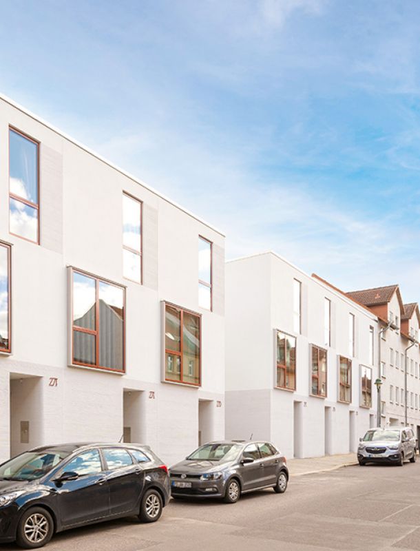 Moderne Stadthäuser mit weißer Fassade, großen Fenstern und urbanem Design in einer lebendigen Wohngegend mit geparkten Autos.