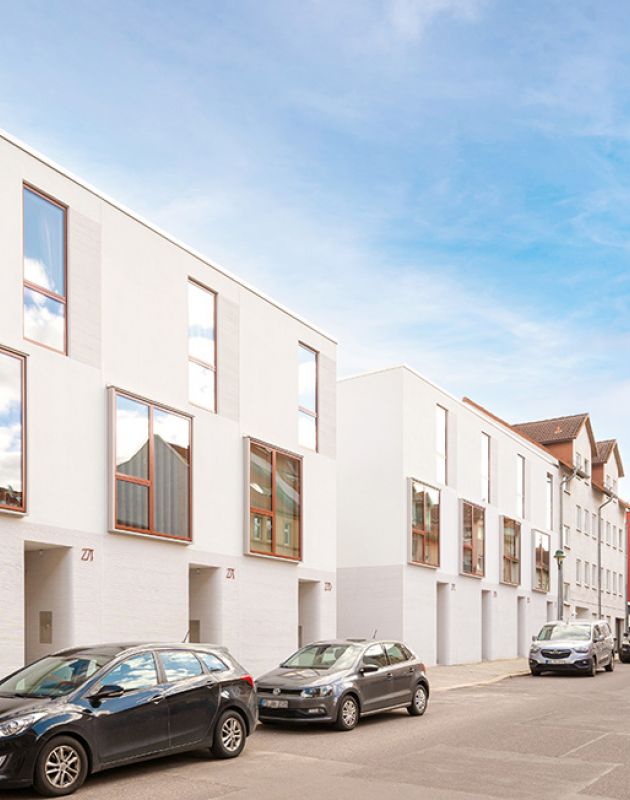 Moderne Stadthäuser mit weißer Fassade, großen Fenstern und urbanem Design in einer lebendigen Wohngegend mit geparkten Autos.