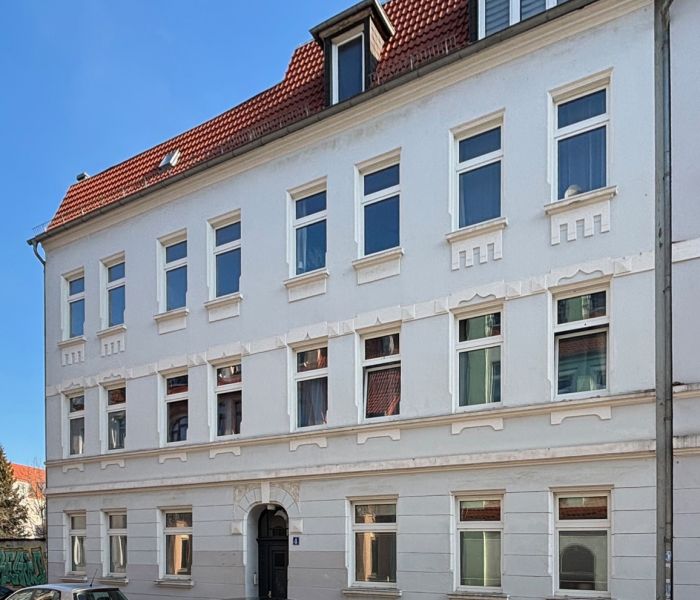Sanierter Altbau mit weißer Fassade und rotem Dach in einer ruhigen Stadtstraße mit geparkten Autos.