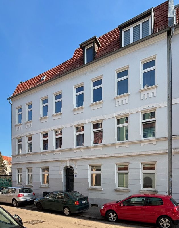 anierter Altbau mit weißer Fassade und rotem Dach in einer ruhigen Stadtstraße mit geparkten Autos.