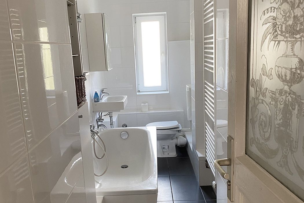 Modernes, helles Badezimmer mit Badewanne, Waschbecken, WC und Fenster, gefliest in Weiß und Schwarz.