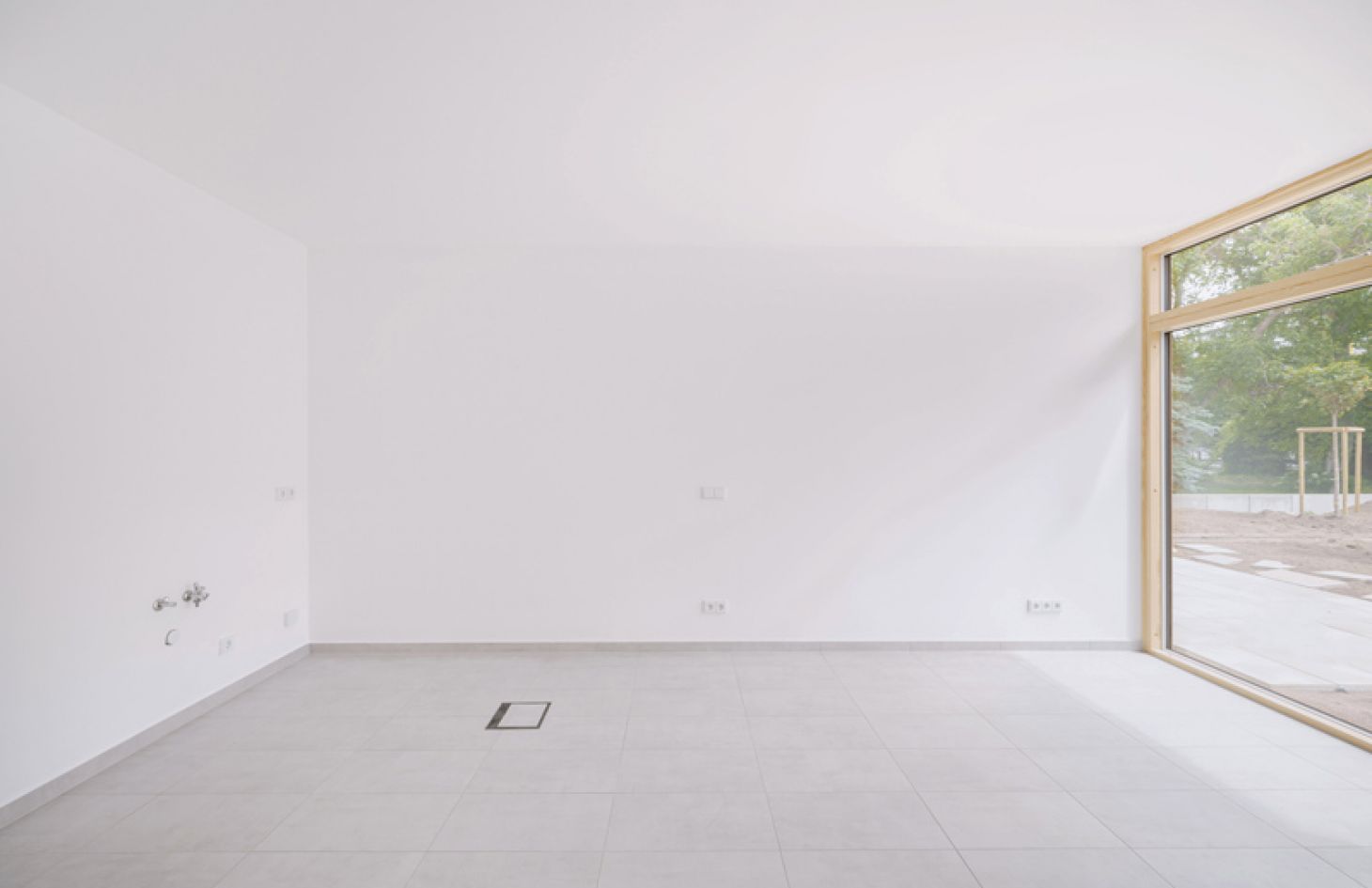 Minimalistischer Wohnraum mit großen Fenstern, weißen Wänden und Fliesenboden, vorbereitet für eine offene Küche mit Blick ins Grüne.