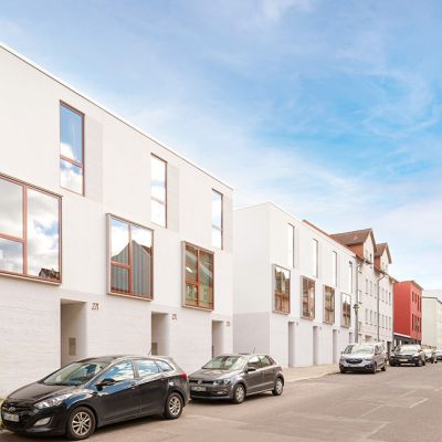 Moderne Stadthäuser mit weißer Fassade, großen Fenstern und urbanem Design in einer lebendigen Wohngegend mit geparkten Autos.